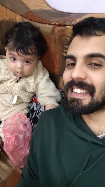 A selfie captures a man and a baby. The man, with dark hair and a beard, wears a green hoodie and smiles. The baby, with dark curly hair, wears a beige fluffy jumpsuit. They sit on a patterned cushion in front of a couch. A pink plastic bag rests near the baby. The background is slightly out of focus.