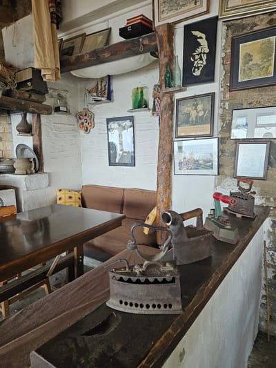 This interior image depicts a cultural or historical establishment in Tire, Turkey. In the immediate foreground, a dark wooden countertop displays a collection of five antique clothes irons of varying designs, including cast-iron models with wooden handles and one with a red handle, all exhibiting signs of rust and age. To the left, a polished dark wooden dining table with a visible chair leg occupies the foreground.
The midground features a brown upholstered bench or sofa positioned against the textured, white-washed walls. A yellow crocheted cushion with sunflower motifs rests on the sofa. The walls are adorned with numerous framed artworks and photographs. Notable items include a black framed artwork with a gold profile of a man's face, a framed black and white photograph captioned "Tire Bandosu 'Amerika yolcusu'" depicting a group of men with musical instruments, and a framed landscape drawing dated "Tire - 1984." Other framed images include portraits, a group photo, and a document. A small wooden sailing ship model hangs from a shelf, and two green glass bottles are placed on another small wooden shelf. A decorative evil eye wall hanging is visible on the wall.
To the far left, a white-washed built-in structure, possibly a fireplace or oven, serves as a display area for a dark pottery vase, a decorative metal plate, and a wooden mortar and pestle. Above this, a vintage radio receiver sits on a wooden shelf next to a woven basket and draped beige fabric. Exposed dark wooden beams and a vertical wooden post are prominent architectural features, contrasting with the rough, white-washed plaster walls. The floor is dark and indistinct. The scene is illuminated by ambient light, suggesting an indoor setting with a rustic, historical ambiance.