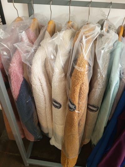 This image presents a close-up, indoor view of multiple winter jackets, each individually encased in a clear plastic garment bag, hanging on wooden hangers with metal hooks. The hangers are suspended from a horizontal bar of a grey metal clothing rack. The jackets are primarily textured fleece, appearing in assorted colors including pink (with a dark blue lower panel), cream/off-white, and mustard yellow. Several garments clearly display a black, circular label with white text, reading "AUGUST WEAR". Additional partial views of jackets in light blue, dark blue, and purple are visible on the right. The setting is in Baza, Myanmar (Burma). No individuals are present.