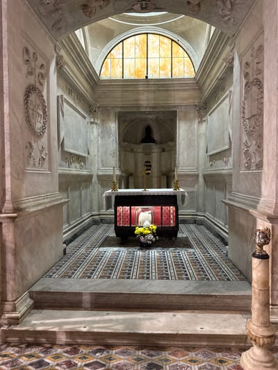 The image depicts the interior of a religious shrine, likely a chapel or a tomb, within the city of Naples, Italy. The architecture is grand and ornate, constructed primarily of marble. A central altar is visible, draped with a white cloth and adorned with a crucifix and two candlesticks. Beneath the altar, a glass-encased relic is displayed, highlighted by a red fabric lining and illuminated from within. A bouquet of yellow and white flowers rests at the front of the altar.
The floor is paved with intricate geometric mosaic tiles in shades of yellow, white, gray, and red. The walls are decorated with bas-relief carvings, including circular wreaths containing inscriptions and figural scenes. An arched stained-glass window with amber-colored panes is positioned at the top of the shrine, allowing natural light to filter in. The overall impression is one of solemnity and reverence, characteristic of a sacred space.