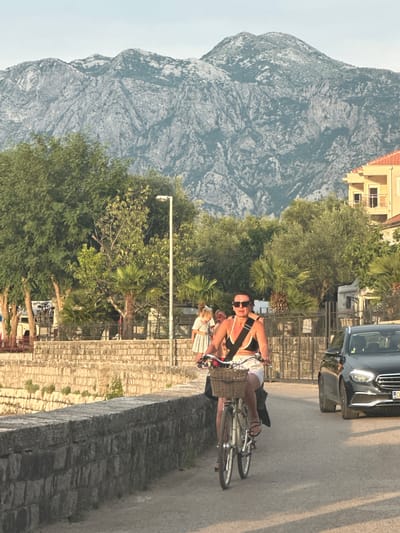 The image captures a scene in Prčanj, Montenegro, likely during a sunny day with clear skies. A person is riding a bicycle on a paved road next to a stone wall. They are wearing sunglasses and a black and white top. Behind them, a dark car is parked on the road. The background features a majestic, rocky mountain with some trees and vegetation on its lower slopes. A building with a red roof is visible on the mountain. The overall atmosphere seems relaxed and pleasant, suggesting a casual outdoor activity. The lighting indicates it is likely late afternoon or early evening, given the warm tones and shadows. The stone wall appears to be a retaining wall or part of a promenade, and the presence of palm trees suggests a Mediterranean climate. There is a street lamp visible, and the road surface suggests it is a public thoroughfare.