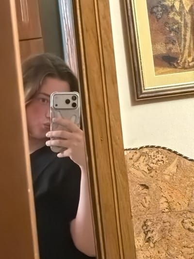 A person is taking a selfie in a mirror. The person is holding a smartphone in their right hand. They are wearing a black t-shirt. The mirror is framed by a wooden border. In the background, there is a framed painting on the wall and a textured surface that appears to be wood or stone. The image is taken indoors, likely in a home or personal space. The exact location in the city of Stryi, Ukraine, cannot be determined from the visual information provided.