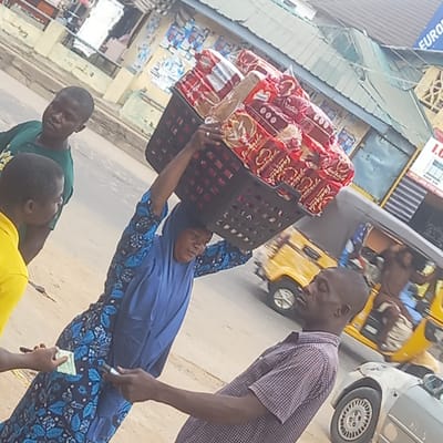 This image captures a vibrant street scene, likely a market or commercial area, in Sardauna, Nigeria, during daylight hours. The central focus is a woman who skillfully balances a large, black plastic basket filled with packaged bread on her head. She wears a blue head covering (hijab) and a patterned blue dress, with her arms raised to steady the heavy load.
She appears to be a street vendor, interacting with customers. A man in a red and white checkered shirt leans towards her, possibly engaging in a transaction or conversation. In the foreground, two younger individuals are visible. One, wearing a green shirt, looks towards the camera, while another in a yellow shirt is seen with his hand extended, holding what appears to be money, suggesting a purchase from the woman. A small currency note is visible in the woman's hand.
The bread packages, predominantly red, clearly display the brand name "Fantastica" and possibly "Fast food." In the background, typical urban elements include a yellow auto-rickshaw (keke Napep) with occupants and a silver car, indicating a busy thoroughfare. Buildings line the street, with partial views of signs. One sign has "EURO" written on it, and another shows "I.G." followed by "OFFICE HOURS." The overall scene depicts a common daily life interaction in a bustling urban environment.