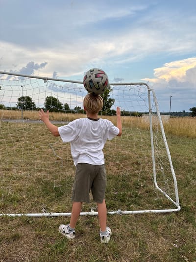 In Bönnigheim, Germany, a young boy stands on dry grass facing a soccer goal. He is wearing a white t-shirt and green shorts, with athletic shoes and gray socks. A soccer ball rests balanced on the crown of his head. His arms are raised slightly to his sides, with his left hand extended towards the goal net. The sky above is partly cloudy with hues of gray and light yellow. The background shows a field of dry grass, trees, and a road. The soccer goal has a white frame and a white net.