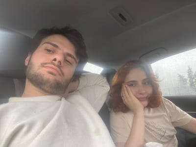 A couple is visible inside a vehicle. The female has auburn hair and is wearing a light-colored t-shirt. She has a ring on her finger and is partially covering her face with her hand. The male has dark hair and a beard, wearing a white sweater. Raindrops are visible on the windshield outside. The location is Unknown, Georgia.