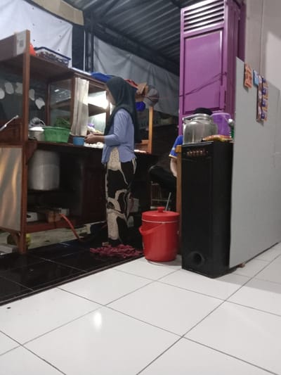 A woman in a blue shirt and black and white patterned pants stands behind a wooden food cart with a large metal pot and a green plastic container on the counter. The food cart is situated in a small room with a purple door, a large black speaker, a red bucket, and a white refrigerator. The room has tiled floors, and there is a partially visible man sitting on a chair in the background. The location is Kecamatan Brangsong, Indonesia, which is identified by the coordinates -6.944 latitude and 110.221 longitude.