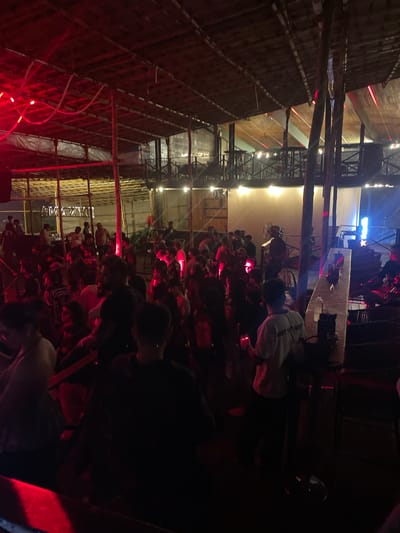 The image depicts a crowded indoor venue, likely a bar or nightclub, in Vagator, India, illuminated by red and white lights. A diverse group of people are present, many standing and socializing. The architecture features exposed wooden beams and a thatched roof, suggesting an open-air or semi-open-air design common in beachside establishments. In the foreground, several individuals are visible, with some appearing to be dancing or engaged in conversation. The background shows more people, tables, and what appears to be a bar area. The overall atmosphere is lively and social.