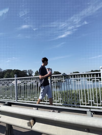 A young man walks along a bridge in Myrhorod, Ukraine, on a sunny day. He is wearing a dark blue polo shirt and light blue denim shorts, with a dark crossbody bag slung over his shoulder. He holds a red object, possibly a beverage container, in his right hand. The bridge has a white metal railing and is elevated over a body of water surrounded by lush green trees and reeds. The sky is a clear blue with a few wispy white clouds. The perspective suggests the photo was taken from another vehicle or a slightly lower vantage point on the bridge.