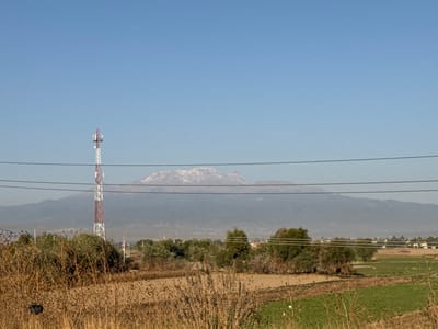 The scene depicts an expansive outdoor environment in Santa Ana Xalmimilulco, Mexico, under a clear, bright blue sky. In the immediate foreground, dry, light brown fields and overgrown, straw-colored vegetation dominate, partially obscuring the ground. Four dark, horizontal lines, consistent with utility wires, stretch across the middle third of the frame. To the left, a tall, lattice-structured cellular tower, painted in alternating red and white sections, rises prominently, featuring multiple antenna arrays near its apex. Beyond the foreground vegetation, segments of green cultivated fields are visible, interspersed with clusters of green trees and shrubs. In the distant midground, a low-density collection of light-colored, single-story structures suggests a settlement. The background is dominated by a large mountain range, its upper peaks partially covered with snow or ice, appearing somewhat hazy against the horizon. No people or animals are discernible, and no active interactions or specific actions are depicted within the static landscape. The overall lighting suggests daylight hours with direct sunlight.