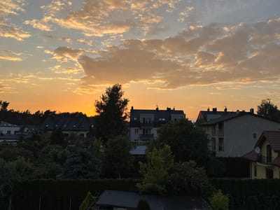 The image captures a residential area in Marki, Poland, during a sunset. The sky is filled with warm hues of orange and yellow, with scattered clouds tinged by the setting sun. In the foreground, lush green trees and hedges dominate the landscape, interspersed with the dark roofs of buildings and the dark surfaces of other structures. Several houses and apartment buildings are visible, some with multiple stories and balconies. The architectural style appears to be typical of suburban European residential areas, with pitched roofs and varied facade colors. The scene conveys a sense of calm and domesticity as the day ends.