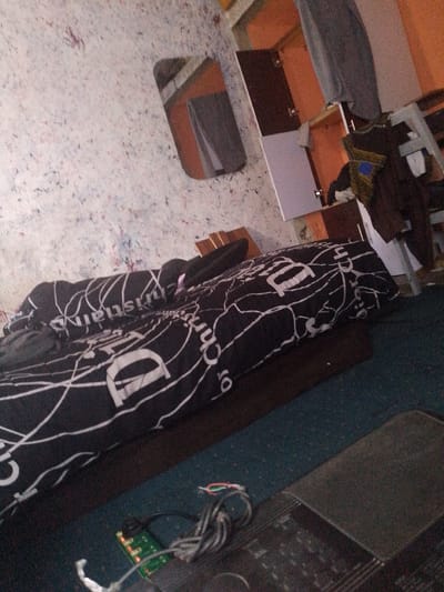 The image depicts a dimly lit bedroom, likely in a residential dwelling in Jimeta, Nigeria. The primary focus is a bed covered with a black comforter featuring a white, abstract pattern with what appears to be lettering. The comforter is rumpled, suggesting recent occupation or disarray. To the left of the bed, the wall is covered in a textured wallpaper with a light, speckled pattern. In the background, there is a wooden wardrobe with orange doors and a mirrored surface reflecting a grey garment. Various personal items and furniture are visible in the background, including what seems to be a shelf unit and possibly a suitcase or bag. On the floor in the foreground, a small green circuit board with wires is partially visible, connected to a black electronic device, possibly a charger or a small appliance. The overall impression is one of a lived-in space, with an unmade bed and the presence of electronic components suggesting active use of the room.