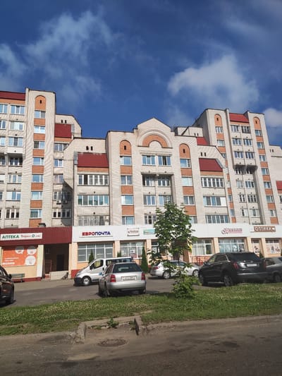 The image captures a street view in Polatsk, Belarus, featuring a modern apartment building with a light brick facade and red tiled roofs. The building has multiple stories and numerous windows. In front of the building, a row of shops is visible, indicated by signs for "АПТЕКА Добрыя лекі" (Pharmacy Kind Medicines), "ЕВРОПОЧТА" (Euro почта - a postal service), and "ОПТИКА" (Optics). Several cars are parked along the street and in a parking lot in front of the shops. The sky is blue with scattered white clouds. The ground is paved, with a strip of green grass and clover in front of the parking area.