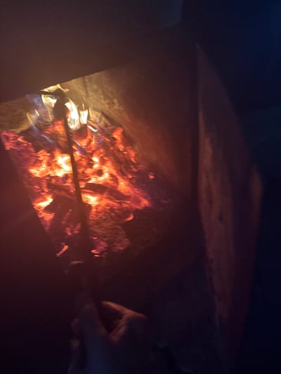 The image shows a close-up view of a fire burning in what appears to be a makeshift fire pit or brazier. The flames are a vibrant orange and yellow, casting a warm glow on the surrounding surfaces. The fire is contained within a dark, metallic structure, the sides of which are visible. A hand, partially obscured, is holding a dark-colored rod or skewer, seemingly inserted into the fire. The background is dark, with some indistinct shapes that could be part of the environment or objects. The overall impression is one of a person tending a fire, possibly for cooking or warmth. There is no specific indication of location within the city of Курган, Russia.