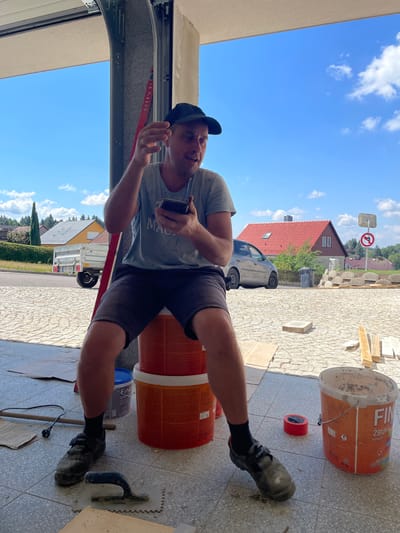 A man wearing a blue t-shirt, dark shorts, black socks, and dark work shoes sits on top of two orange buckets and a blue bucket. He is wearing a black baseball cap and holds a dark-colored smartphone in his right hand, with his left hand raised as if gesturing. The man is in the foreground, in what appears to be a driveway or garage. Behind him, a silver car is parked on the street, and a house with a red roof is visible. The background shows a bright blue sky with some clouds and green trees. Construction materials, including wood planks and bags of what appear to be cement or other building materials, are scattered around the scene. A red level tool leans against a pillar to the left of the man, and a trowel and other tools are on the ground near his feet. The location is identified as the city of Jablonné nad Orlicí, Czechia.