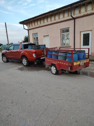 In Babuk, Bulgaria, a rusted red trailer is filled with seven blue barrels and is hitched to an orange Ford Ranger pickup truck parked on a paved road. The truck and trailer are positioned in front of a pale pink building with weathered windows and ornate architectural details above the windows. The sky above is a pale blue with scattered clouds. The scene appears to be outdoors in a rural or semi-rural setting, likely during the daytime.