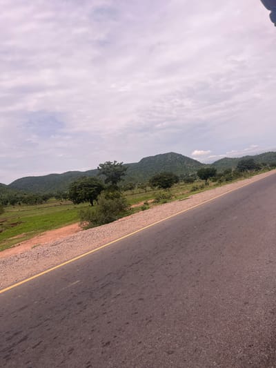 This media file is a landscape photo, taken from the perspective of someone traveling on a paved road. The road, with a yellow line marking its edge, occupies the right side of the frame and extends diagonally into the distance. To the left of the road, there's a strip of gravelly earth, followed by a vibrant green field that stretches towards a backdrop of lush, tree-covered hills. The hills are undulating and covered in dense vegetation, indicating a natural, possibly rural or undeveloped area. The sky above is partly cloudy, with streaks of white and grey clouds against a lighter sky, suggesting daytime. There are no people or specific objects visible, creating a sense of solitude and focus on the natural scenery. The overall impression is one of travel through a scenic, possibly tropical or subtropical, environment. The location context provided is Minda, Nigeria, which aligns with the visual cues of green hills and vegetation. The image appears to be taken during daylight hours, with no specific time of day being evident beyond that. The weather seems fair, with clouds present but not obscuring the sun. There is no visible text in the image.
