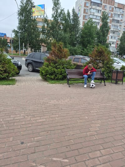 The image captures a street scene in Nyagan, Russia, with a young person sitting on a park bench next to a soccer ball, engrossed in their smartphone. The bench is made of dark wood and metal, and the soccer ball is black and white. The background features a multi-story building with balconies, trees, and parked cars, including a black SUV and a lighter-colored sedan. The ground is paved with red bricks. A digital billboard displaying colorful graphics and Russian text is visible above the trees in the distance. The sky is overcast.