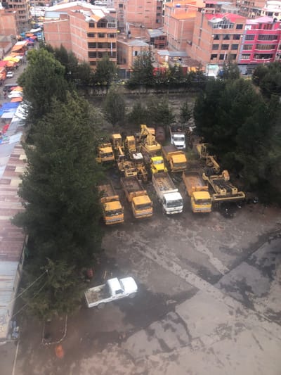 An aerial perspective reveals a dense urban landscape, juxtaposed with a construction vehicle storage area. Brick buildings, tinged with red hues, form a stark contrast to the gray, concrete ground. Trees dot the scene, partially obscuring the view of a collection of yellow bulldozers, trucks, and a white pickup truck. Laundry lines laden with colorful clothes suggest residential life interspersed with industrial activity.