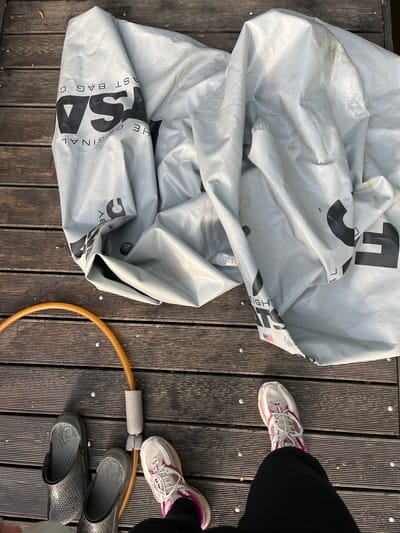 The image shows a section of a weathered wooden deck. On the left side, a large, deflated, light gray vinyl bag with the words "THE ORIGINAL LAST BAG CO." printed in black is crumpled. To the right of the bag, a person is standing with their feet visible. They are wearing running shoes (white and pink) and black slip-on shoes (gray). Between the shoes is a section of orange hula hoop that connects with a gray foam cushion. The location is not possible to know.