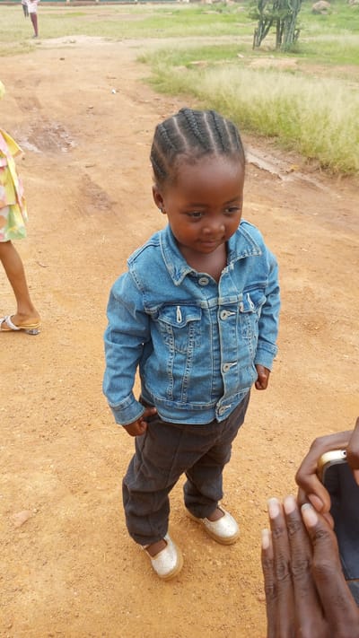 A young child with braided hair stands on a dirt surface in what appears to be an outdoor environment, possibly near Barikin Sabon Gidan Kanat, Nigeria. The child wears a denim jacket, dark trousers, and light-colored shoes. Another person's legs and feet are visible on the left side of the frame, while a hand holding a smartphone is on the right. The background features sparse vegetation and a building in the distance.
