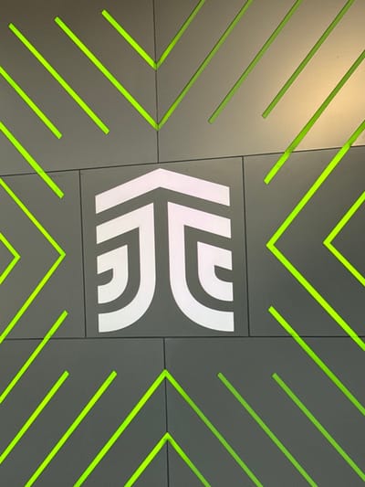 An indoor wall segment features a modern, geometric design. The surface consists of large, rectangular dark gray panels arranged in a grid. Centered on these panels is a distinct white, illuminated logo, composed of thick, rounded lines forming a stylized upward-pointing arrow or combined initialism, consistent with the Metrobús León (Sistema Integrado de Transporte - SIT) logo. Surrounding this central emblem, and extending across the entire visible wall, are numerous integrated neon green linear elements. These green lines are arranged diagonally, forming a repeating pattern of V-shapes and parallel strokes, acting as illuminated grooves within the panel structure. No individuals or actions are depicted. The scene is located within a station of the Metrobús León system in León, Guanajuato, Mexico.