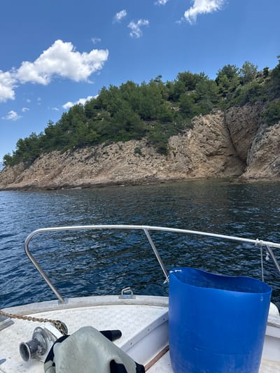 The image shows a view from the bow of a boat sailing on a body of water. In the foreground, part of the boat's deck is visible, including a metal railing, a winch with a chain, and a large blue plastic container. The water is dark blue with ripples. In the background, a rocky, tree-covered hillside rises steeply from the water's edge. The sky above is blue with scattered white clouds. This scene is consistent with the coastline near Potos, Greece, a seaside town located on the southern coast of the island of Thasos.