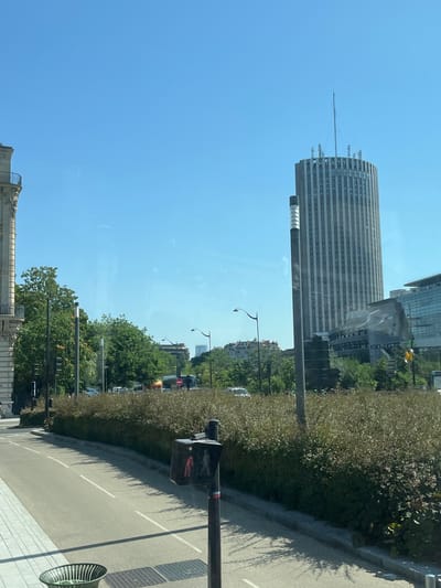 In the city of Neuilly-sur-Seine, France, a street scene is captured on a clear day with a bright blue sky. On the right, a tall, cylindrical skyscraper with vertical grey stripes and a radio antenna at its peak dominates the skyline. The letters "H Y A T T" are visible near the top of the building. To the left of the skyscraper, a modern, glass-faced building is partially visible. In the foreground, a paved road with white lane markings curves to the left. A pedestrian signal displaying a red walking figure is present on the right side of the road. Dense green bushes line the median strip separating the road from the buildings. Trees are scattered throughout the scene, some taller and more mature, others shorter and bushier. A traffic light and street lamps are also visible along the road.