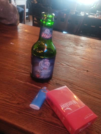 A bottle of "Royale" beer, a blue lighter, and a pack of Winston cigarettes rest on a wooden table. The bottle has a blue label with a lion logo and Arabic text. The scene appears to be indoors, possibly in a bar or restaurant, given the wooden furniture and ambient lighting. The table's surface is made of planks with visible grain. The background is blurry, showing hints of other patrons and furniture, suggesting a social setting. The lighting is dim with a bright light source at the top, creating reflections on the bottle. The overall composition is from a slightly elevated angle, focusing on the objects on the table. The location in Essaouira, Morocco, is not definitively indicated by any visual cues in the image itself.