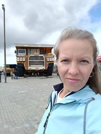 This is a medium shot selfie taken outdoors, featuring a woman in the foreground and a large mining truck in the background. The woman, who appears to be in her late 30s or early 40s, has light skin, blue eyes, and shoulder-length grey-blonde hair. She is wearing a light blue hooded jacket with a dark collar over a white top. She has hoop earrings in both ears. The woman is smiling slightly and looking at the camera.

Behind her, a colossal yellow mining truck is parked on display. The truck is enormous, with massive tires and a large cabin. Its front grille and headlights are visible, as well as part of its cargo bed. The truck is elevated on platforms, suggesting it is a museum piece or monument. To the left of the main truck, another, smaller dark blue truck is partially visible.

The setting is an outdoor plaza paved with grey interlocking stones. A tall lamppost stands to the left of the frame. In the distance, a few other people can be seen, appearing as small figures relative to the trucks. The sky is cloudy with patches of blue.

The visible text on the truck, oriented horizontally and likely in Cyrillic script, reads: "ВЕРХНЕПЫШМИНСКИЙ КОМПЛЕКС" which translates to "VERKHNEPYSHMINSKY COMPLEX" and "ОТЦА" which is likely part of a longer word or phrase. There is also some text on the woman's jacket, appearing to be a brand name in a stylized font.

The overall impression is of a visit to an industrial or mining museum in Verkhnyaya Pyshma, Russia, on a day with mixed weather. The woman appears happy and engaged with her surroundings.