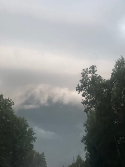 The image shows a view of the sky with dramatic, dark clouds gathering, suggesting an impending storm. Silhouetted trees frame the lower portion of the image, with dense foliage visible on the left and right sides, and a line of trees extending into the distance at the bottom. The sky is overcast, with lighter clouds forming a band across the upper part of the frame, and darker, more ominous clouds filling the lower sky. A faint, possibly reddish, circular light or anomaly is visible in the upper center of the sky. The scene appears to be outdoors in a natural setting, possibly a rural area or the outskirts of a town, given the presence of trees and the open sky. The location is identified as Полянка Снт (контемирово), Russia. There are no people or animals visible in the image. The overall atmosphere is somber and potentially foreboding due to the weather conditions.