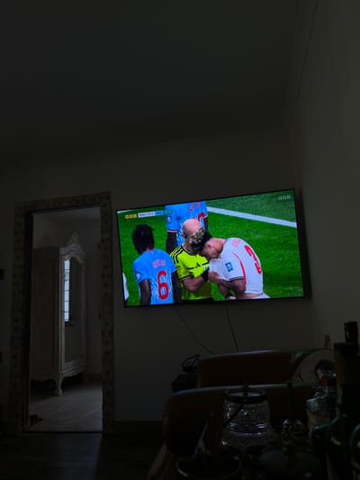 The media file shows a television screen displaying a soccer match. Two players are prominently featured, with one in a white jersey and the other in a yellow jersey, appearing to be in a physical exchange on the field. The background shows a green grass field with white lines, typical of a soccer pitch. The television is mounted on a wall, and below it, a large mirror with an ornate frame is visible, reflecting parts of the room. The overall scene suggests an indoor setting, likely a living room or entertainment area, where someone is watching the soccer game. The lighting is dim, indicating it might be evening or the room is not well-lit.
Visible text on the screen includes "BBC" in the top left corner, a scoreline that reads "DRC 0 | 1 ENG", and "48:46", indicating the time elapsed in the match. Another player's name, "BYAU", is visible on the back of a blue jersey with the number "6". The reflection in the mirror shows a dark interior with some furniture and possibly other reflective surfaces. The presence of a television and mirror in a home environment is the primary context for this media. The emotion displayed by the players on the screen appears to be one of intense physical engagement, common in sports.
