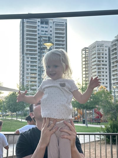This is a portrait photograph, taken outdoors, of a young blonde girl held by an adult, likely her father. The girl is looking at the camera with a smile, her right arm outstretched. She is wearing a white top with small pink dots and light pink pants. The adult is supporting her with their hands. Behind them, two modern apartment buildings are visible. One building has a large gold, horn-like structure on its facade. Trees and a playground with red equipment are also in the background, suggesting a residential area. The time of day appears to be late afternoon or early evening, indicated by the warm, soft sunlight. The weather seems pleasant and clear. The overall mood is one of familial affection and joy.