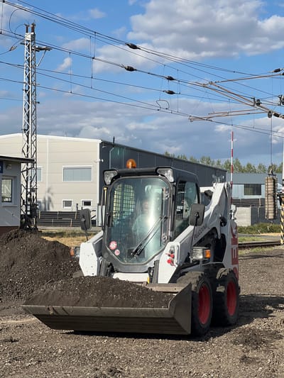 In Újezd u Brna, Czechia, a Bobcat compact track loader, identifiable by the text "PŮJČOVNA" (rental) on its side, is actively moving a pile of dark soil. The loader's bucket is filled with the soil, suggesting an ongoing construction or landscaping project. In the background, a light gray industrial building with several windows is visible, along with railway tracks and overhead power lines, indicative of the area's infrastructure. A red and white striped barrier arm, commonly seen at railway crossings, is also present. The sky is partly cloudy, suggesting a daytime scene. There is a person visible inside the cab of the loader.