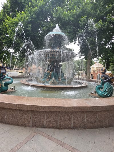 The image displays a decorative fountain with water jets spraying upwards and outwards, creating a dynamic visual effect. The fountain is multi-tiered and appears to be made of stone and metal, with sculpted figures resembling mermaids or mythical creatures incorporated into its design. The water cascades into a pool at the base.

In the background, lush green trees provide a natural backdrop, with their leaves filling a significant portion of the frame. Behind the trees, a building with arches and an ornate facade is visible, suggesting an urban or park-like setting. The ground around the fountain is paved with stone tiles.

The activity taking place is the operation of the fountain, with water actively flowing and spraying. The scene is likely set during the daytime, given the bright lighting and the clarity of the details. The weather appears pleasant, as the fountain is operating and the trees are in full foliage.

Notable details include the intricate design of the fountain's sculptures and the textural contrast between the smooth water, the rough stone of the fountain and paving, and the soft leaves of the trees. The overall impression is one of a public space designed for aesthetic appeal and enjoyment. There is no visible text in the image. The location context provided is Ganja, Azerbaijan.