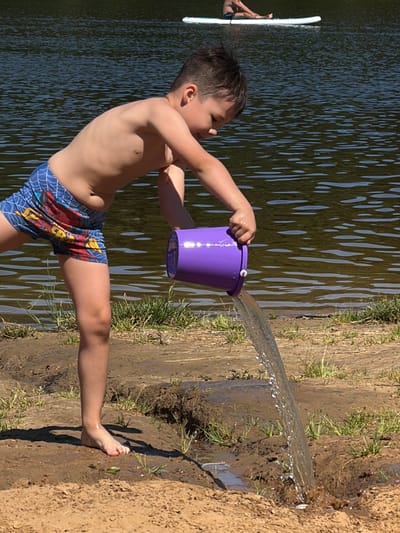 A young boy, shirtless and wearing blue swim trunks with a spider-web pattern and a red and yellow superhero print, stands on the sandy bank of a body of water. He is holding a purple bucket in his right hand, tilting it to pour water onto the ground. A stream of water is actively flowing from the bucket. His left arm is extended slightly, and his body is angled towards the water. The boy's face is partially visible, and he appears to be concentrating on his task.
The background consists of dark, rippling water, suggesting a lake or pond, with sunlight reflecting off its surface. On the right side of the frame, the sandy bank is wet and muddy where the water from the bucket has landed, with sparse green grass growing along the edge. The time of day appears to be sunny, indicated by the bright lighting and the shadows cast on the ground. The overall scene depicts a child playing with water on a warm day, likely in a natural outdoor setting like a park or beach, possibly in Malakhovka, Russia, given the location context. There is no visible text in the image.