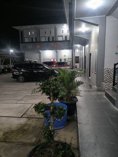 The image depicts an outdoor scene at night, likely a parking area or pathway next to a building complex. A black SUV is parked on a paved surface composed of large, square concrete slabs. To the left of the SUV, two motorcycles are parked. In the foreground, two potted plants are visible, one a tall palm and the other a smaller, more dense shrub. The building to the right features a covered walkway with a grey tiled floor and a light grey exterior wall with multiple windows and doors. The building has multiple stories, with visible lighting on the upper levels. The sky is dark, indicating it is nighttime. Based on the provided context, the scene is located in the city of Panteraya, Indonesia. There are no people visible in the image.