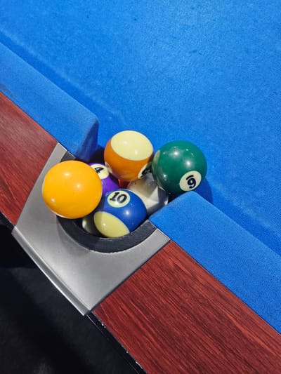 The image presents a close-up, high-angle view of a corner pocket on a billiard table, situated in Arnhem, Netherlands. The table is surfaced with bright blue felt and features matching blue cushions. A segment of the table's dark reddish-brown wooden frame is visible on the left side, bordered by a metallic, silver-toned pocket liner. Six billiard balls are clustered within and immediately adjacent to the pocket. A solid yellow ball rests on the inner lip of the pocket. To its right, a partially visible purple striped ball shows the numeral '4'. Below the purple ball, a blue striped ball displaying the numeral '10' is visible. An orange striped ball is partially observable above the purple ball. A solid green ball, marked with the numeral '9', rests on the blue cushion just outside the pocket. A white ball is partially visible behind the blue '10' ball. No individuals are present in the frame. The scene is illuminated by what appears to be overhead lighting.