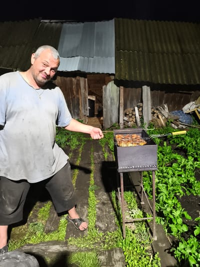 A man with graying hair and a beard stands in front of a grill outdoors at night. He is wearing a light blue t-shirt and dark shorts. He is holding the handle of the grill with his right hand, which contains pieces of meat. The grill is situated on a metal stand in a garden area. Behind him is a wooden structure that appears to be a shed or outbuilding, with a corrugated roof. Piles of firewood are visible to the right of the structure. The ground is covered with wooden planks and greenery. The overall setting suggests a rural or suburban environment in Il'mino, Russia.