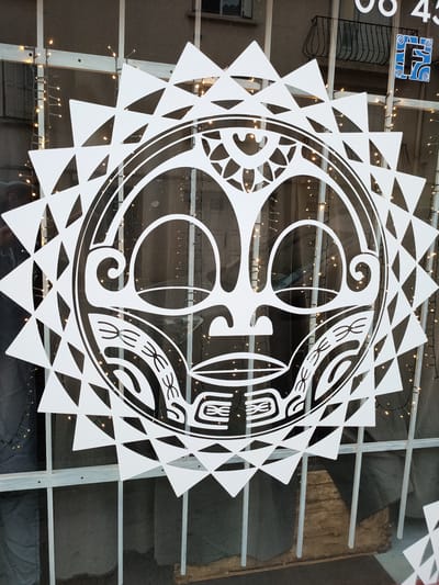 This is a close-up, eye-level, medium shot of a large white, sun-like graphic adhered to a storefront window. The graphic features a central face with stylized Polynesian-inspired motifs, surrounded by triangular points. Behind the graphic, a string of small, warm white fairy lights is visible, draped across a window frame and partially obscured by the graphic. The window itself has a light grey frame and vertical bars. In the upper right corner of the image, a blue and white logo with the letter "F" is partially visible, along with part of a dark numerical display that reads "08 43". A black wrought iron balcony is visible through the window in the background, as is a dark, indeterminate interior space. The image appears to be taken during daylight hours, possibly in the city of Perpignan, France.