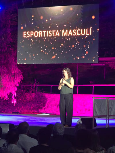 The image shows a woman signing on a stage in front of a large screen at an event in Cornellà de Llobregat, Spain. The screen displays the text "ESPORTISTA MASCULÍ" in large, glowing letters against a dark background with scattered golden orbs. The woman, wearing a dark sleeveless top and black pants, is positioned in the center of the stage, facing slightly to her left. Her hands are raised in a signing gesture. The stage is lit with vibrant pink and blue lights, illuminating the backdrop and casting shadows. In the foreground, the backs of several audience members are visible, seated in darkness. The overall scene suggests an outdoor evening event, possibly an awards ceremony or a cultural performance.