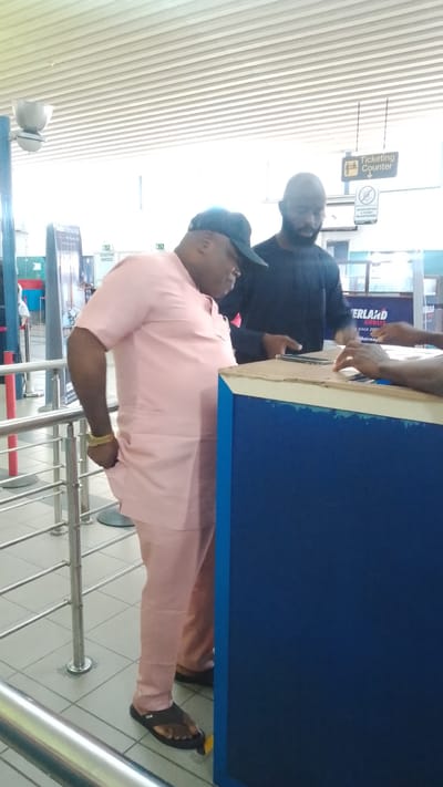 The image depicts two men interacting at a counter, likely in a ticketing office or check-in area, within an airport or transportation hub. The man in the foreground is wearing a light pink traditional Nigerian outfit and a dark baseball cap. He has a gold bracelet on his left wrist and is wearing black flip-flops. The man behind him is wearing a dark long-sleeved shirt and has a beard. They appear to be engaged in a transaction or discussion.
In the background, a sign reads "TICKETING OFFICE" with an arrow pointing down. Another sign below it says "NO EXTORTION TICKET RACKET RING". Part of a flight information board is visible with "ERLAND AIRWAYS" and text below it. The environment appears to be indoors with tiled flooring, metal railings, and a suspended ceiling with horizontal panels. The lighting is bright, suggesting daylight or strong artificial lighting. Based on the context of an airport and signage, this scene could be located at the ticketing counter of an airport in Orerokpe, Nigeria, or a similar transportation facility.
