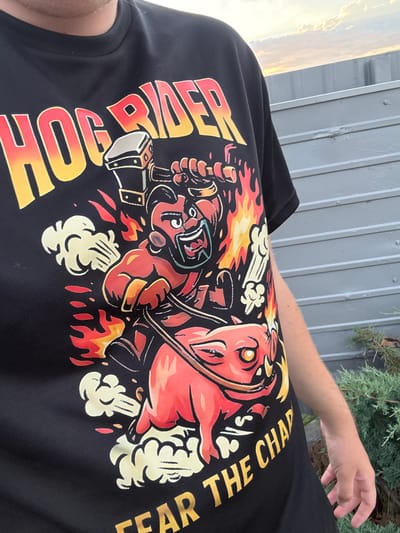 A person wearing a black t-shirt with a "Hog Rider" graphic is pictured outdoors. The graphic depicts a cartoonish figure riding a red, fiery pig, with flames and smoke elements surrounding them. The text "HOG RIDER" is prominently displayed at the top of the shirt in yellow and red gradient, and "FEAR THE CHAP" is visible at the bottom. The background shows a grey, corrugated metal fence and some green foliage. The sky above is partially cloudy with hints of sunset colors. The image appears to be taken during daylight. The location is not specified to be Zatoka, Ukraine.