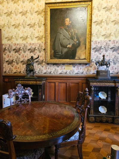 The image shows an interior view, likely a room in a historical building or museum, possibly located in the Алупкинский городской совет, Ukraine. On the left, a framed portrait of a man in a dark coat hangs on a wall with floral wallpaper. Below the portrait is a bronze statue of a figure pointing upward. Adjacent to the statue and portrait is a dark wooden panel, possibly part of a built-in cabinet or wall feature. On the right, there's a large, round wooden table with ornate details. Part of a chair is visible beside the table, indicating this is a dining or meeting space. The floor is made of wooden parquet with a geometric pattern. The scene is richly decorated, suggesting a historical period.