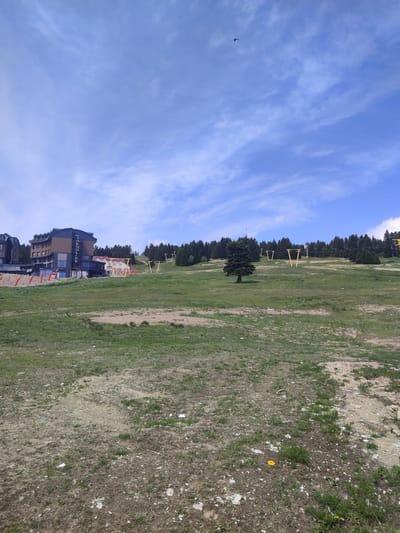 An outdoor, ground-level, wide shot captures a grassy slope leading up to a line of trees under a bright blue sky with scattered white clouds. In the distance, yellow ski lift towers are visible among the trees. To the left of the frame, a multi-story hotel building with dark and light brown facades stands prominently, with its name "TRENDLIFE" vertically displayed on its side. Orange safety barriers are arranged in a line in front of the hotel. The foreground consists of a patch of dry, uneven ground with sparse green grass and scattered white debris. A single dark bird is visible in flight against the sky.