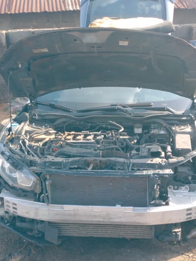 A close-up view of the front section of a dark-colored car is presented, with its hood fully open. The car's front bumper is removed, exposing the internal components of the engine bay and the forward chassis. A prominent silver-colored impact bar is visible across the front, with a large radiator directly behind it and a smaller heat exchanger (likely an intercooler) positioned below.
The engine bay is cluttered with various mechanical and electrical components, including the engine block, numerous hoses, wiring harnesses, a blue-capped coolant reservoir on the left, and a brake fluid reservoir towards the center. A car battery is visible on the right side of the engine bay. The engine area appears dusty and shows signs of wear, indicating regular use or maintenance. On the underside of the open hood, a small red warning label is visible, though its text is too blurry to decipher.
The background consists of a corrugated metal roof structure and a concrete block wall, suggesting an outdoor, possibly workshop or residential, setting. Bright daylight illuminates the scene, creating reflections on the car's windshield where another vehicle's shape is faintly visible. The ground beneath the car is uneven dirt. The overall image depicts a vehicle undergoing repair or inspection, possibly following an incident, in Sardauna, Nigeria.