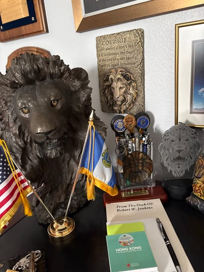 The image shows a desk with various items, predominantly featuring lion motifs. In the foreground, there is a large, dark-colored bust of a lion's head, flanked by two small flags, one of which is the American flag. To the right of the lion bust is a clear container holding numerous pens and lighters, topped with small lion figurines and emblems. Behind this pen holder is a framed plaque with a lion's head design. Further to the right, another lion-themed decorative item is visible. On the desk, in front of the pen holder, are a card that reads "From The Desk Of: Robert W. Jenkins" and a small green booklet with "HONG KONG THE WORLD'S MEETING PLACE" printed on it. A pen rests on top of the booklet. In the background, a framed piece of art with a lion image and motivational text is mounted on the wall, alongside other framed items. The overall setting appears to be an office or personal workspace. There are no people or active interactions depicted in the image. The environment is indoor, with a plain wall and a dark desk surface. The location is identified as Sun City Center, United States of America.