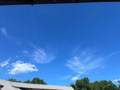 The image captures a clear blue sky with scattered wispy white clouds. In the foreground, a corrugated metal roof is visible, partially obscured by lush green trees along the horizon. A dark overhang, possibly part of a building or structure, runs along the top edge of the image. The scene appears to be an outdoor environment, likely in a semi-urban or rural setting within Ливадийский поселковый совет, Ukraine, given the natural elements and rooftop structures. There are no people or visible actions taking place.