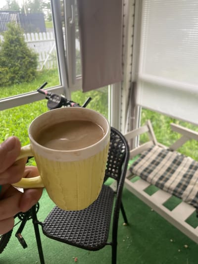 In this media file, a person's hand is holding a yellow textured mug filled with a light brown liquid, presumably coffee. The mug is positioned in the foreground, with its rim slightly tilted towards the viewer. The scene appears to be an outdoor or semi-outdoor setting, possibly a porch or sunroom, given the presence of large windows and what looks like outdoor furniture.
Behind the mug, a black chair with a textured seat is visible. Further in the background, a white bench with a green and black checkered cushion is situated. Outside the window, lush green foliage suggests a natural environment, and a portion of a bicycle is visible, with its handlebars and pedals in frame.
The lighting suggests it is daytime, likely late morning or afternoon, with soft, diffused light. The overall mood conveyed is one of a relaxed, casual moment. While the exact location is given as Bol'shoe Zaborod'e, Russia, the visual cues within the image itself do not provide specific identifiers for this location beyond general outdoor scenery. There is no visible text in the image.