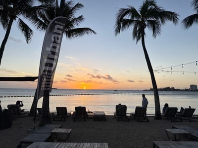 The image displays a sunset over a sandy beach in Noumea, France. The sun, a distinct orange sphere, is positioned low on the horizon over calm ocean water, illuminating the sky with gradients of orange, yellow, and light blue, with scattered wispy clouds.
In the foreground, a sandy beach area contains approximately ten dark lounge chairs, some occupied by individuals facing the sunset. One person on the far left is seated, oriented towards the water, and holding a dark, rectangular object. A man wearing a light-colored collared shirt stands near the water's edge, partially silhouetted, facing the horizon. Multiple tall palm trees are silhouetted against the sky, one of which hosts a prominent grey and white feather flag vertically displaying the word "Urban" multiple times. String lights with visible bulbs are strung between two palm trees on the right side of the frame.
A line of orange or yellow buoys extends horizontally across the water, marking a boundary. In the distant background, a cluster of buildings forms a skyline on the right, while a small boat is visible on the far left of the horizon.