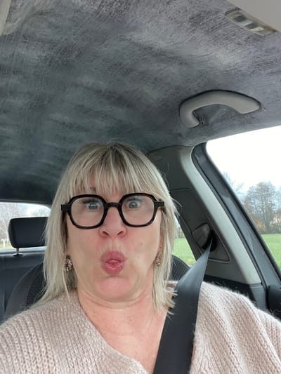 A single adult Caucasian female is depicted from the chest up inside a vehicle, appearing to take a selfie. She has blonde, shoulder-length hair with bangs and wears dark-rimmed hexagonal glasses, patterned earrings, and a light-colored knit sweater. Her facial expression features wide-open eyes and a puckered mouth with the tongue slightly extended. A black shoulder seatbelt diagonally crosses her torso. The vehicle's interior includes a light grey, textured headliner and a grey overhead grab handle on the passenger side. Through the vehicle windows, a blurred exterior environment is visible, characterized by an overcast sky, green fields, and distant bare deciduous trees, indicative of a rural or semi-rural setting. A white signpost with a dark top is partially visible in the far-left background. The image contains no explicit geographical markers to confirm its location in Veauche, France.